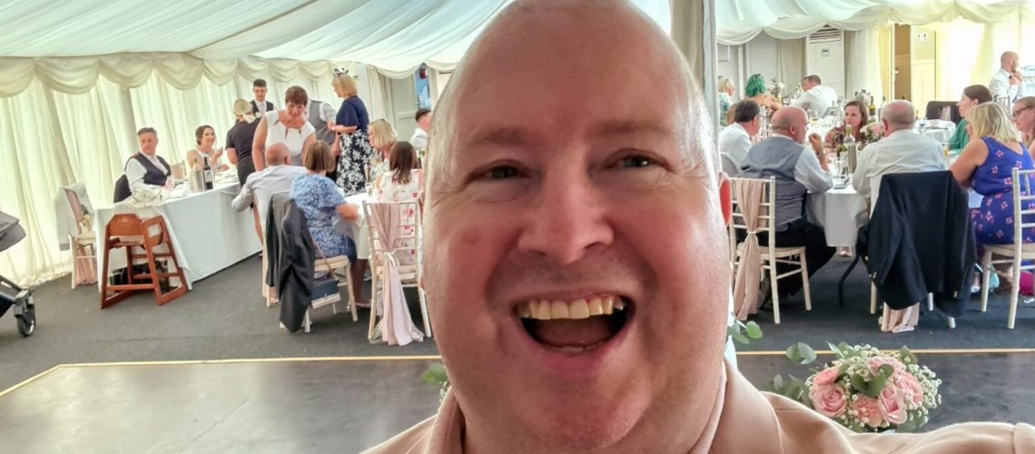 Smiling wedding host/MC in pink suit with seated wedding crowd in background